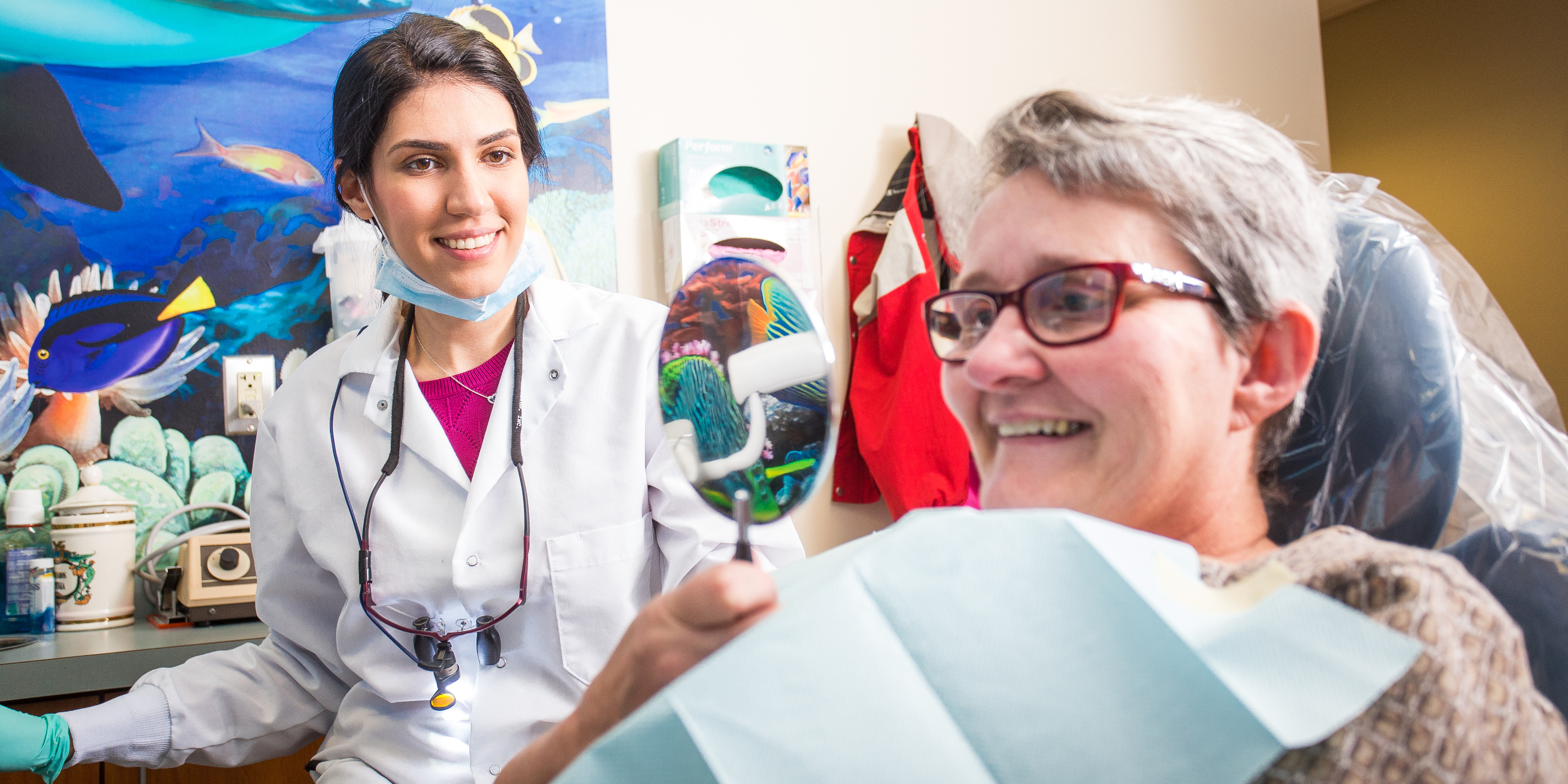 A dentist holds a mirror up to a patient in a chair. Both are smiling.