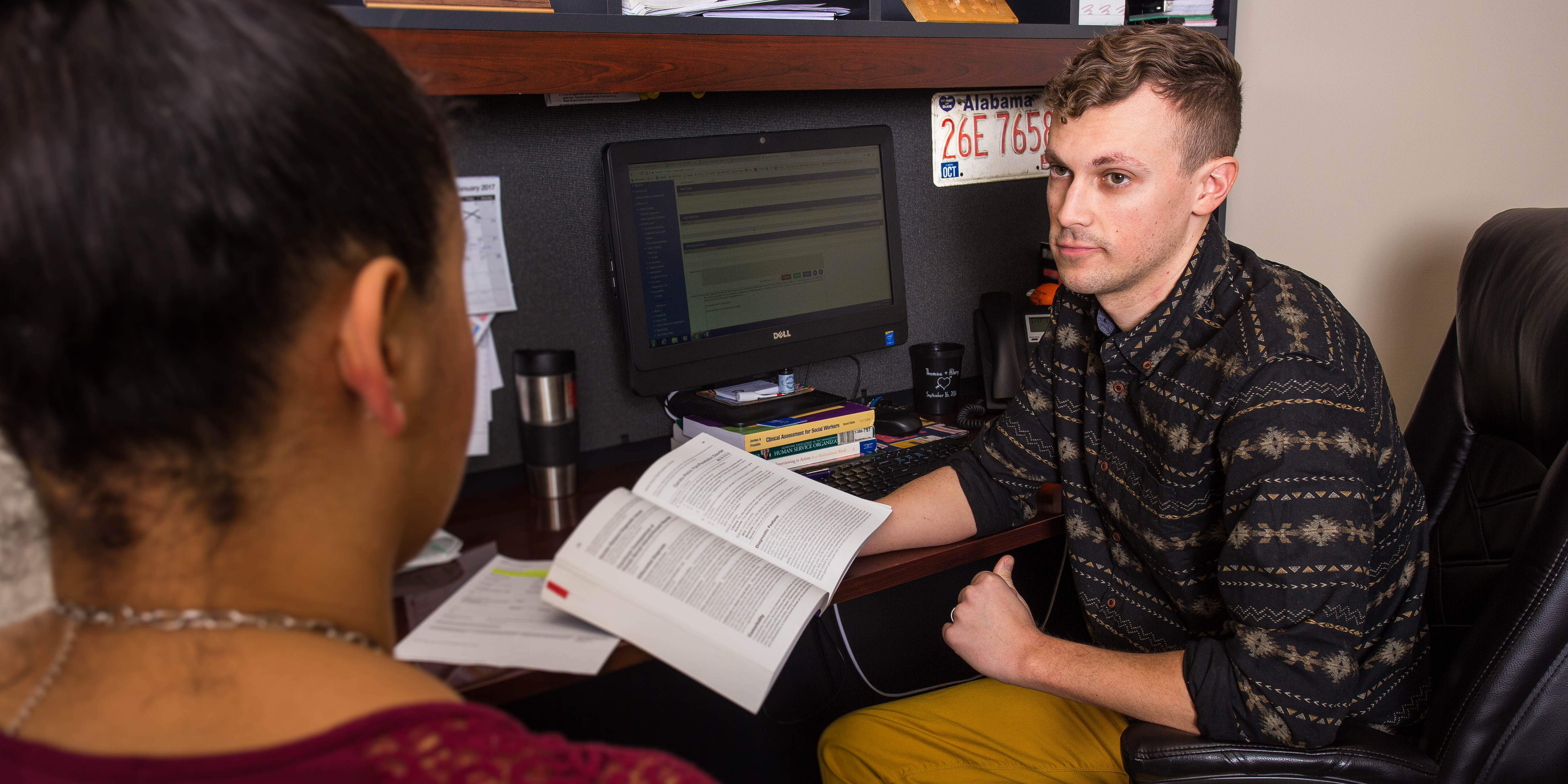 A young man holding a book at a desk looks at a young woman