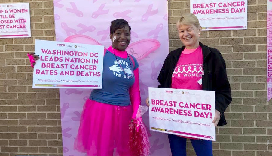 Two women wearing pink clothing standing in front of a brick wall holding breast cancer awareness signs