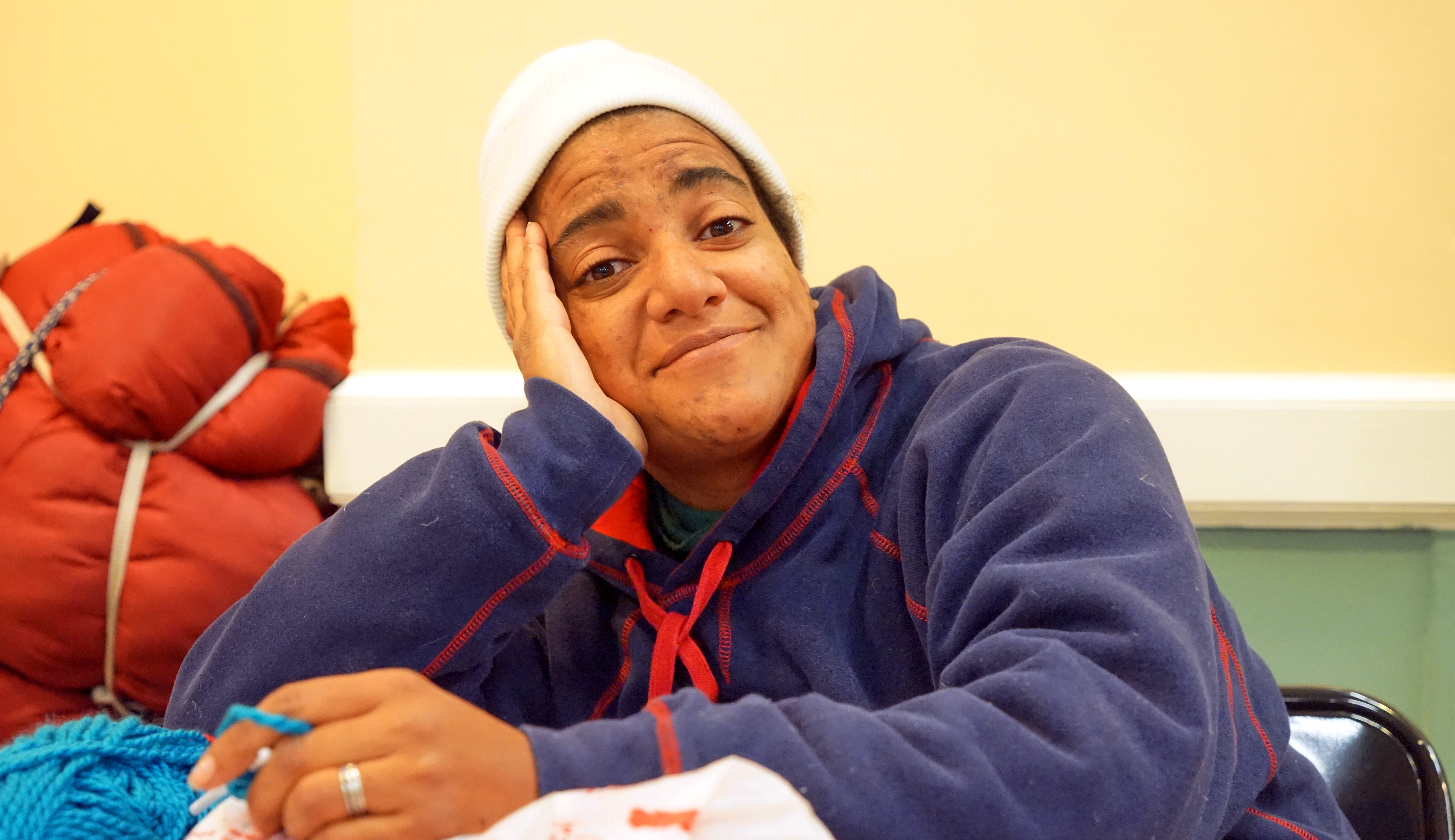 A smiling homeless woman with her elbow propping her head up on a table.