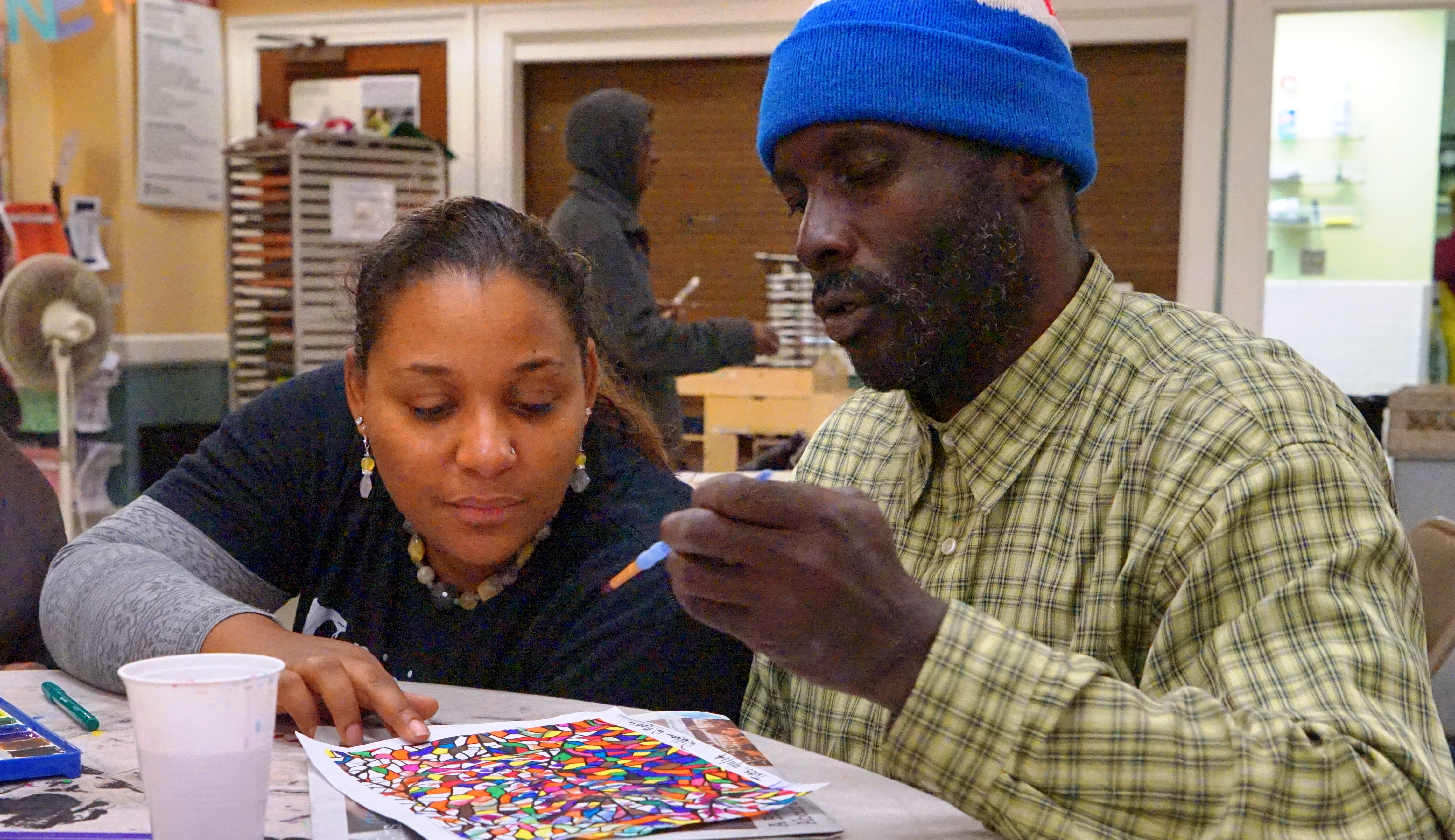 A woman pointing at a piece of artwork on a table that a homeless man is coloring