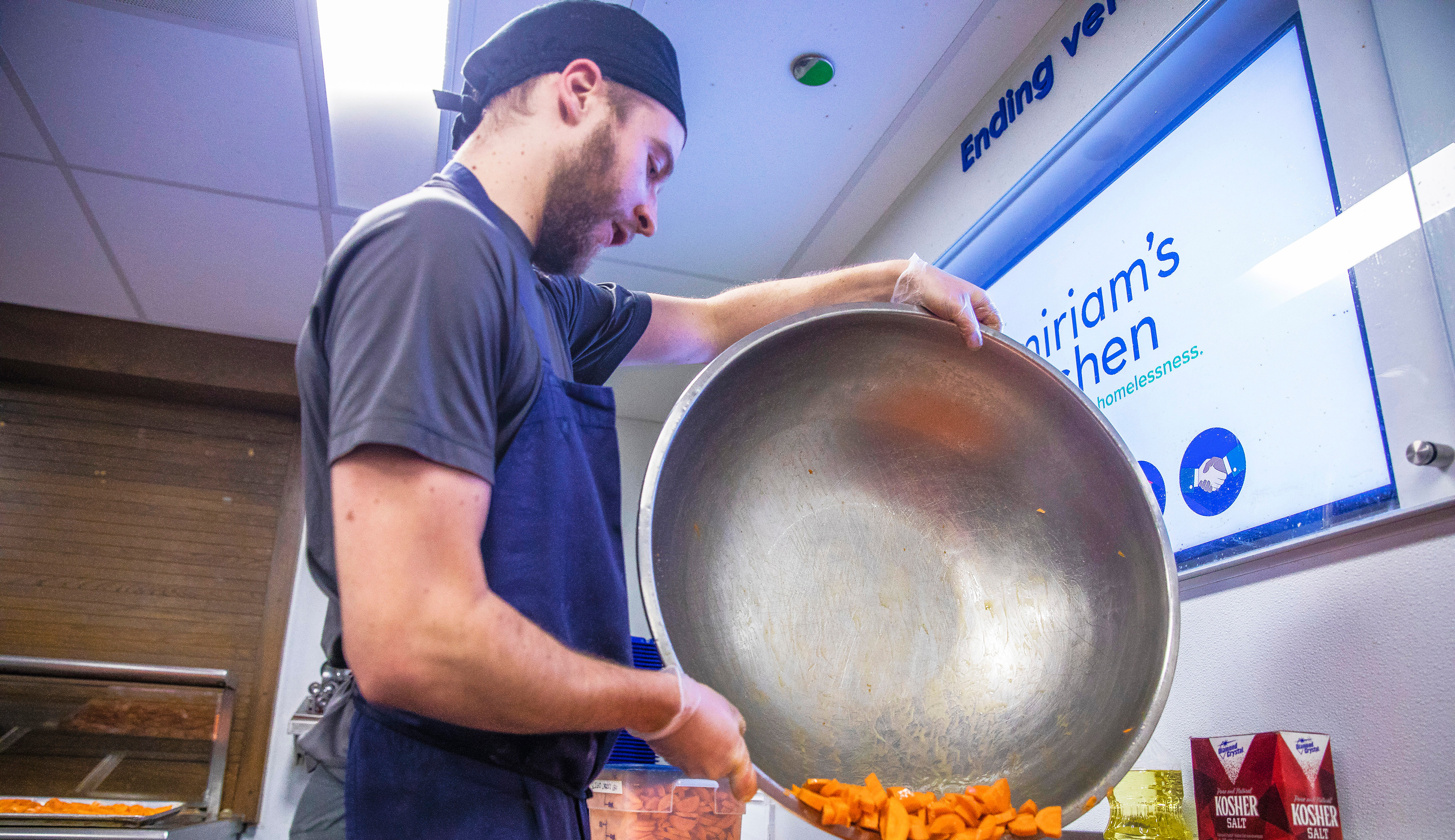 A kitchen worker dumping a large metal bowl of chopped carrots