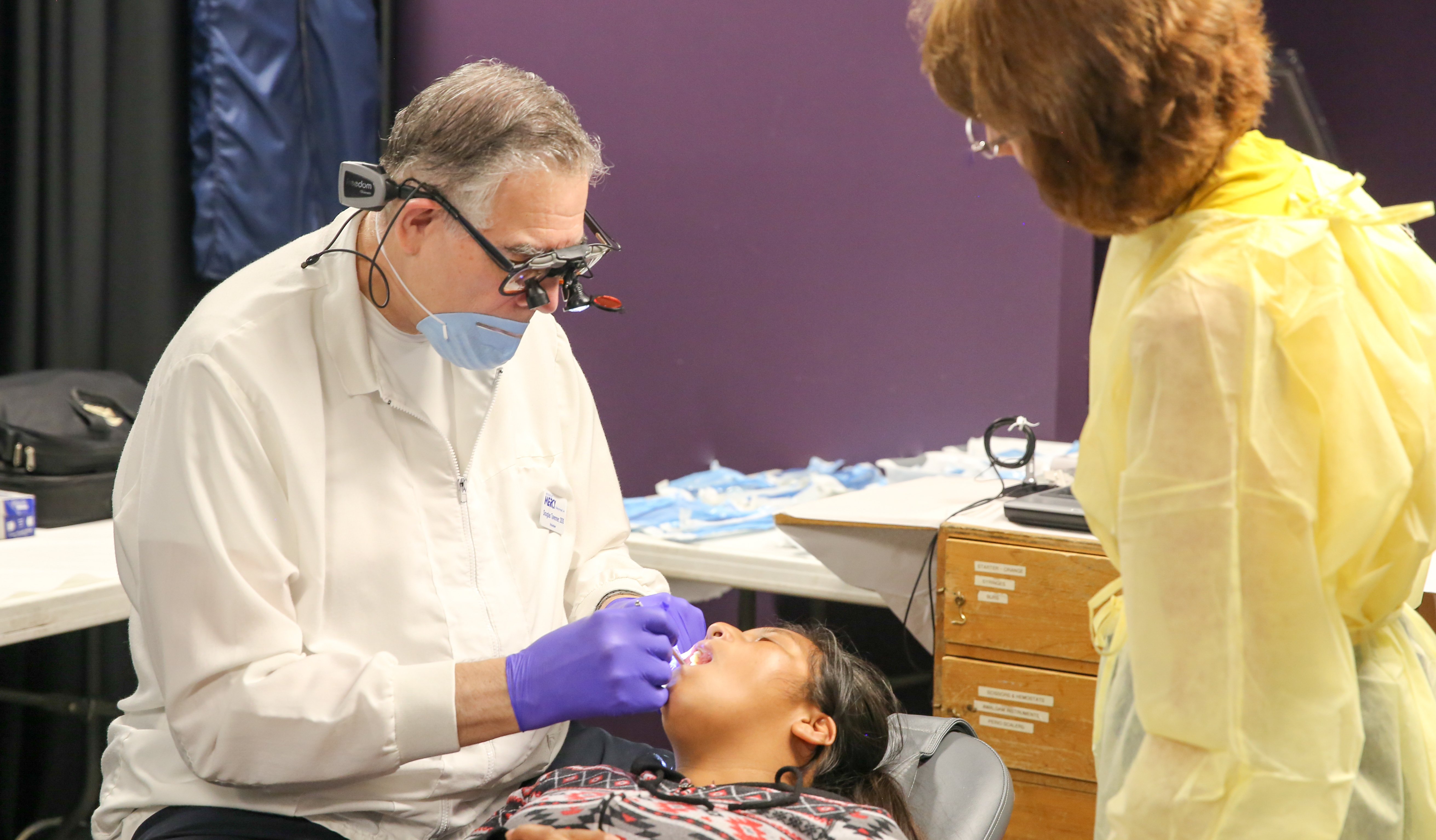 an older dentist examines a patients teeth while a woman looks on