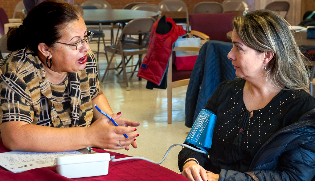 Two women at a table talking while one has their blood pressure checked