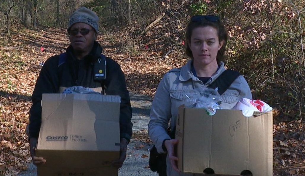 Two people carrying cardboard boxes along a wooded path