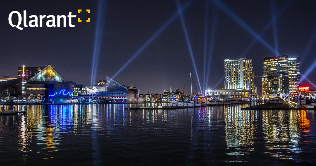 Baltimore Inner Harbor at night with spotlight beams in the sky. Qlarant logo in upper left corner.