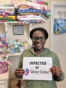 Young, bespectacled, African American woman holding a sign that reads Impacted by Grace Center