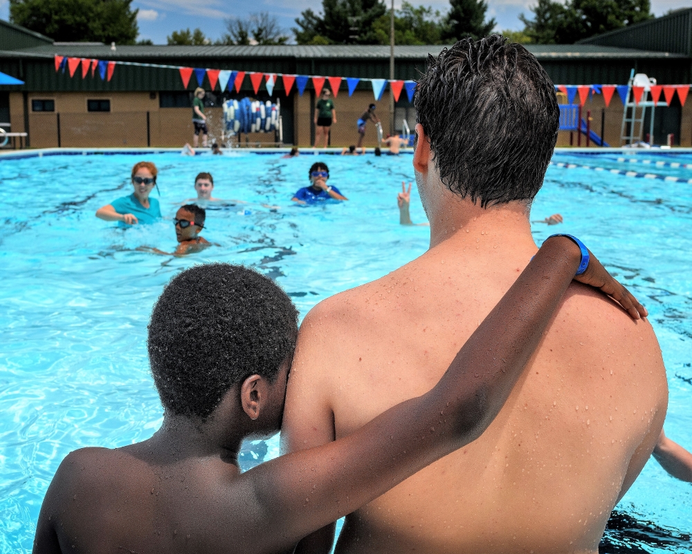 a young african american boy with his arm across the back of a caucasian man - both face away from the camera looking out over a pool of children