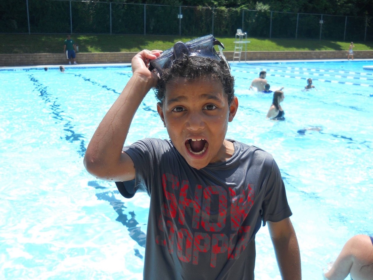 a young boy pulls the goggles off his head in front of a pool