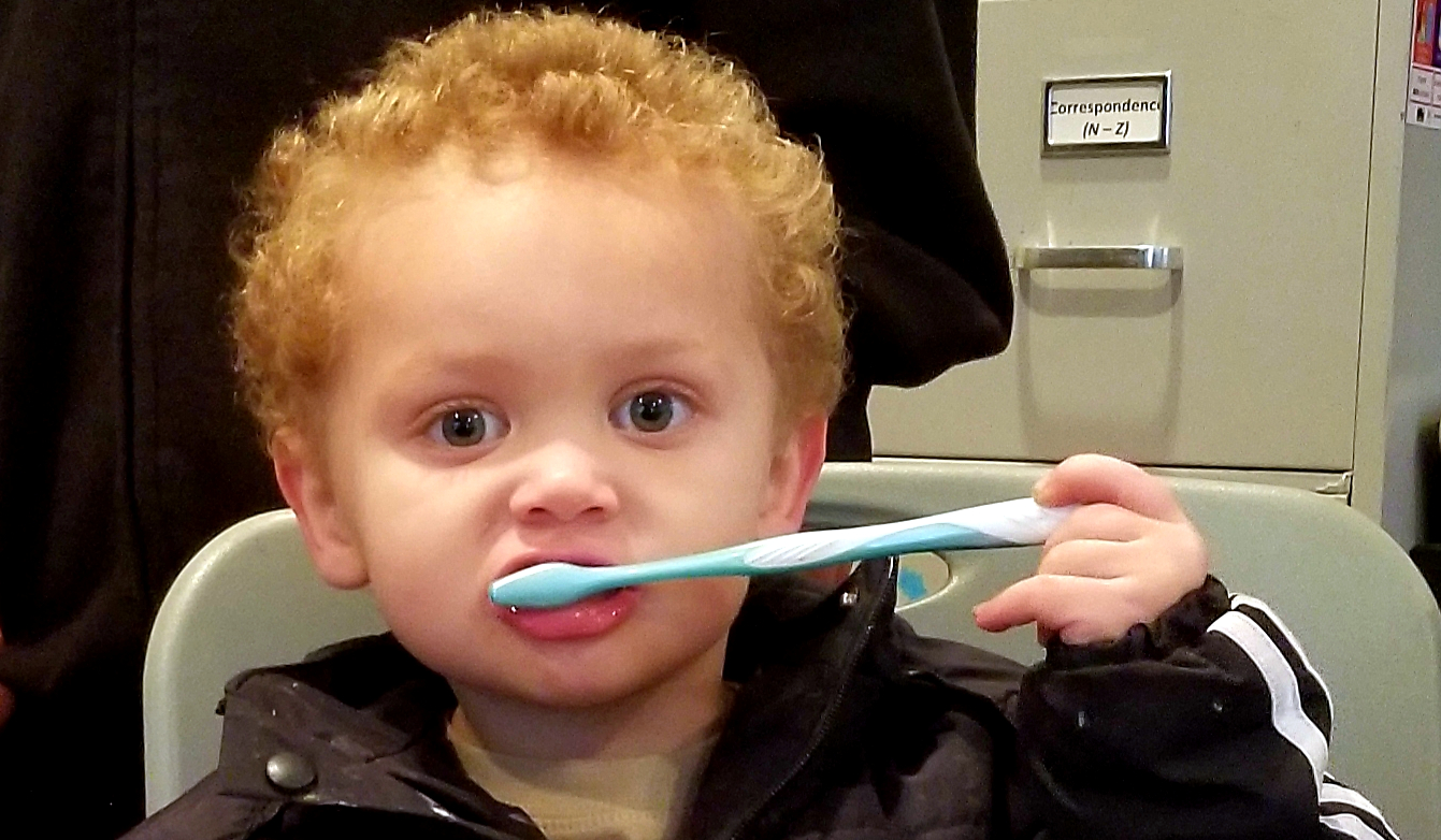 A toddler with curly strawberry blonde hair brushing his teeth