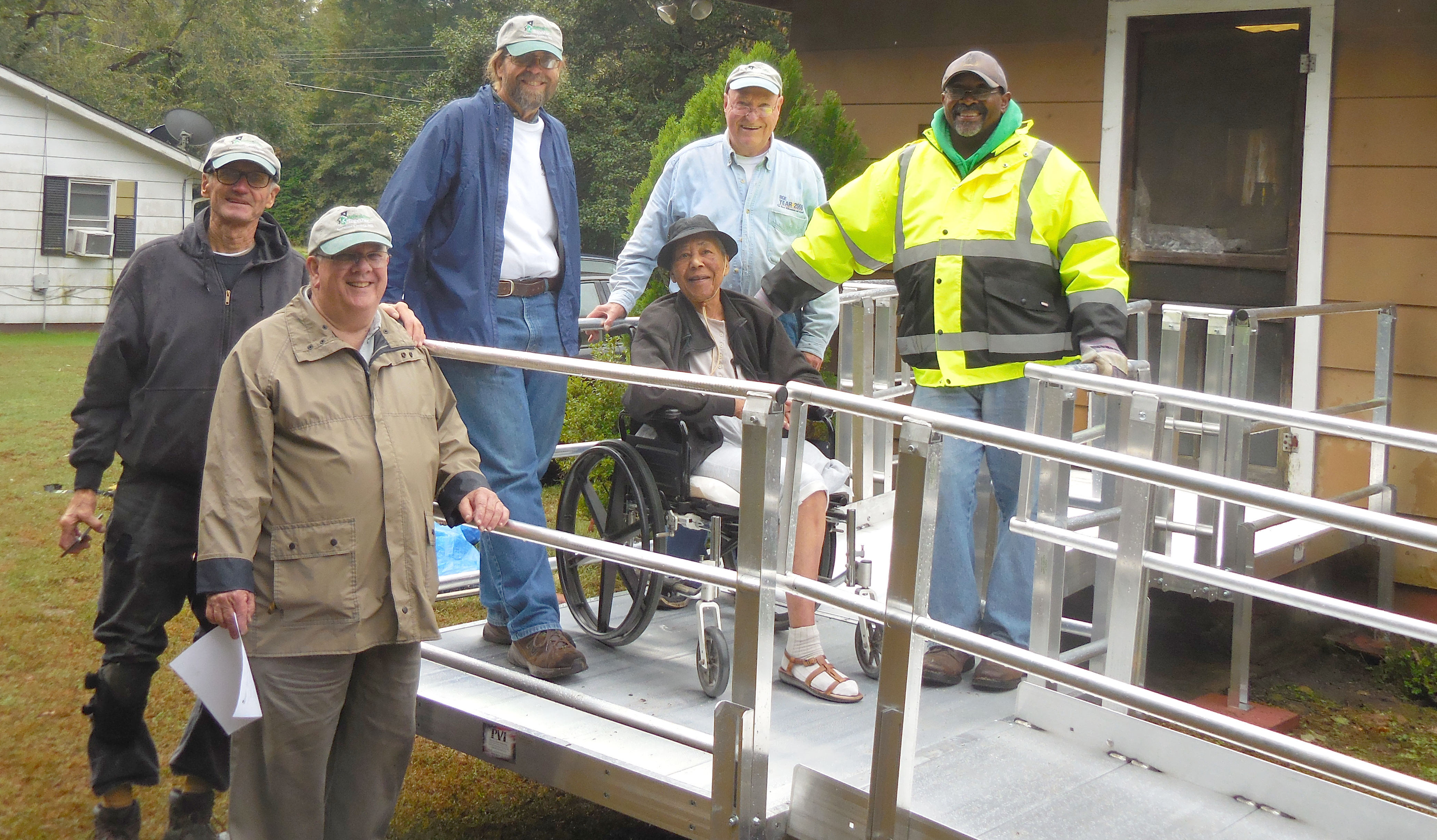 A group of workers stand on a newly built wheelchair ramp with a woman in a wheelchair