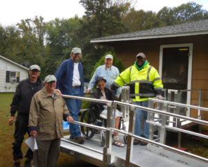 A group of men stand around a woman in a wheelchair on a ramp outside her home