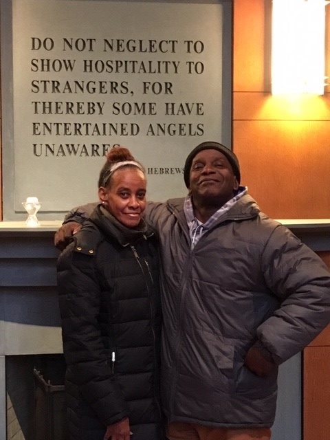 John and Joretta proudly standing together in front of a sign with a quote from the book of Hebrews which reads, Do not neglect to show hospitality to strangers, for thereby some have entertained angels unawares.