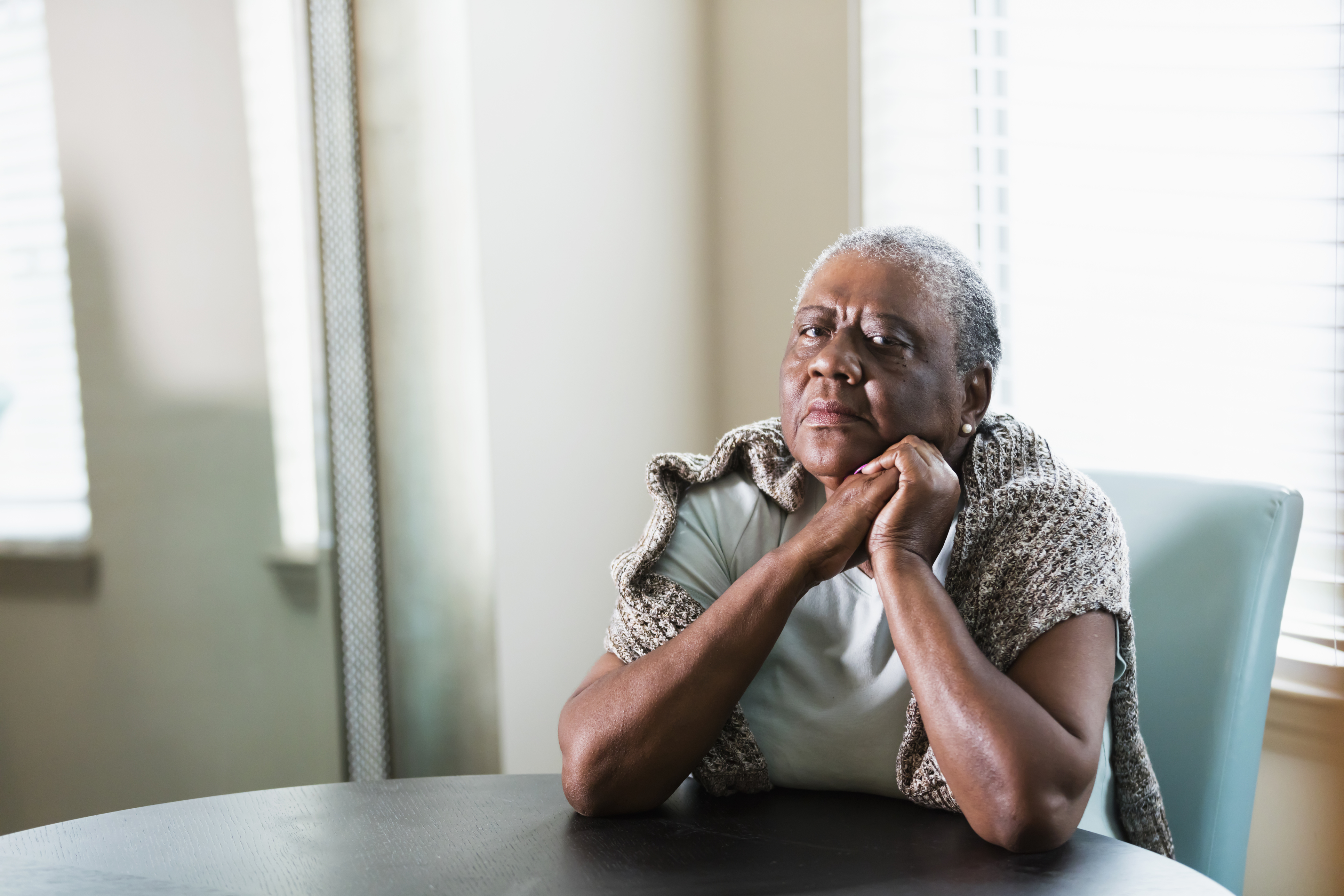 A lonely senior African-American woman in her 70s sitting at home at a table by a window. She is looking at the camera with a serious expression, elbows on the table and hands clasped.