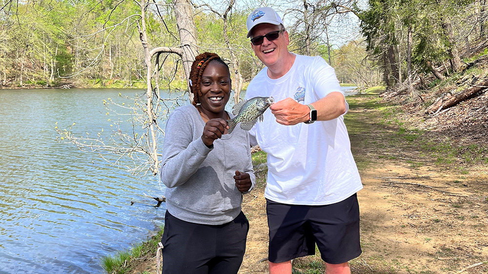 A man and woman hold a fish by a lake